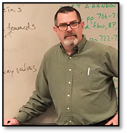 Upper body shot of Stephen Shoemake smiling in front of a whiteboard. Stephen has short dark hair, a graying beard and mustache, and dark eyes.